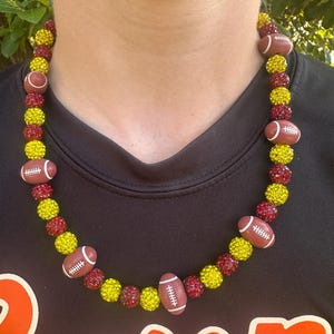Op de afbeelding: Een ketting met voetbalvormige hangers en afwisselend rode en gele kristallen kralen. De ketting wordt om de nek gedragen en is een accessoire met een sportthema.