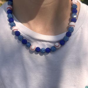 Puede incluir: Un collar de cuentas con temática de béisbol azul y blanco. Las cuentas están hechas de pequeños diamantes de imitación redondos y brillantes. El collar está hecho de cuentas azules y blancas alternas, con un diseño de béisbol en las cuentas blancas.