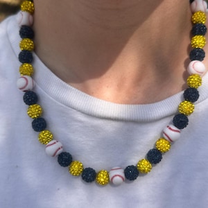 May include: A necklace made of black and yellow beads with white baseballs. The beads are covered in rhinestones.