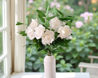 Op de afbeelding: Een boeket witte rozen in een witte keramische vaas. De rozen zijn in een tros gerangschikt met groene bladeren. De vaas is hoog en slank.