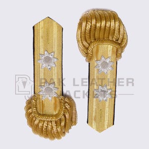 Könnte beinhalten: Paar goldfarbene militärische Schulterstücke mit silbernen Sternen-Emblemen. Die Schulterstücke haben ein strukturiertes, vertikales Streifenmuster und sind mit schwarzem Stoff eingefasst. Die Enden sind mit gewickeltem Goldfaden verziert.