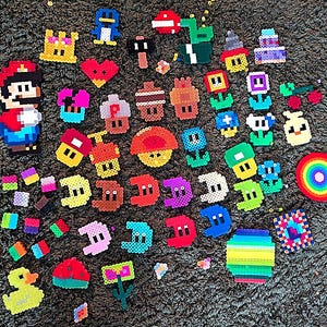 Könnte beinhalten: Eine Sammlung bunter Pixel-Art-Charaktere und -Objekte, darunter Mario, ein Pilz, eine Blume, ein Pinguin, ein Regenbogen und ein Schloss. Die Figuren bestehen aus kleinen Plastikperlen.