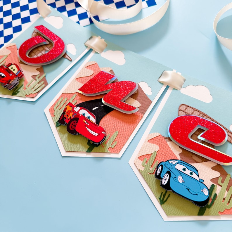 May include: A Cars-themed banner with red, white, and blue accents. The banner features the letters "J", "C", and "E" in red glitter, along with images of Lightning McQueen and Mater.