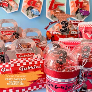 May include: Red and white party favor boxes with the name "Gabriel" printed on them. The boxes are filled with small toys and treats. There are also clear plastic cups with red and white checkered designs and the number "3" printed on them. The cups are filled with red and white sprinkles. The image also includes a banner with the name "Gabriel" and the number "3" printed on it. The theme of the party is Cars.