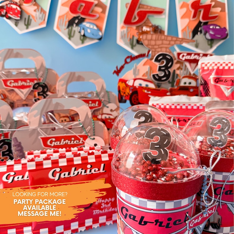 May include: Red and white party favor boxes with the name "Gabriel" printed on them. The boxes are filled with small toys and treats. There are also clear plastic cups with red and white checkered designs and the number "3" printed on them. The cups are filled with red and white sprinkles. The image also includes a banner with the name "Gabriel" and the number "3" printed on it. The theme of the party is Cars.