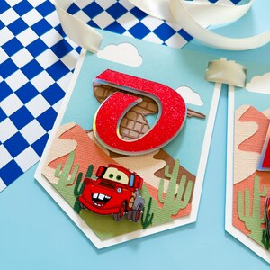 May include: A red and white car with a smiling face is on a banner with a red glitter letter "O" and a desert scene. The banner is blue and has white clouds.