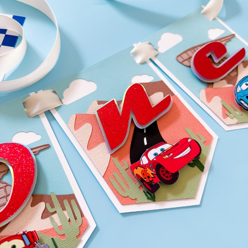 May include: A red, white, and blue Cars-themed birthday banner with red felt letters spelling "ONE" and a red Lightning McQueen car on a desert-themed background.