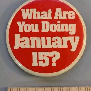 May include: A round red pinback button with white text that reads What Are You Doing January 15? The button is positioned above a ruler showing a diameter of approximately 1.75 inches. The button has a white border and a smooth, glossy finish.