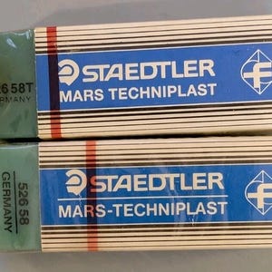 May include: Two rectangular Staedtler Mars Techniplast erasers with green rubber ends and white sleeves featuring blue stripes, black text reading 526 58T and 526 58, the word Germany, and the Staedtler logo. The sleeves have thin black horizontal lines.
