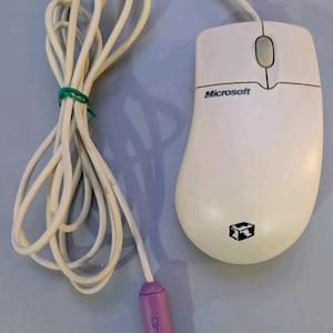 May include: A beige wired computer mouse featuring the Microsoft logo and a black cube icon on the shell. The device includes a long white cord secured with a green twist tie and terminates in a purple PS/2 connector plug at the end of the cable.