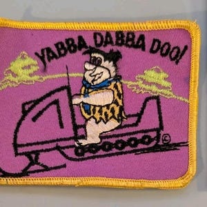 May include: A rectangular embroidered patch with a purple background featuring Fred Flintstone riding a snowmobile. The text YABBA DABBA DOO! is stitched in black above the character. A yellow border surrounds the patch, which measures 3 inches wide.