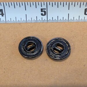 May include: Two round black buttons with a textured outer rim and a central bar, positioned below a white measuring tape that shows the buttons are approximately 0.5 inches in diameter. The buttons feature a dark, matte finish.