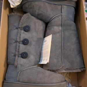 May include: A pair of grey mid-calf boots with three dark circular buttons on the side, featuring a soft interior lining and visible stitching. The boots are placed inside a cardboard box with a white UGG brand tag tucked into the side.
