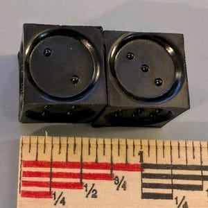 May include: Two matte black cubic dice with recessed circular faces and raised pips, positioned above a wooden ruler showing a measurement of approximately one inch in width for the combined pair of dice.