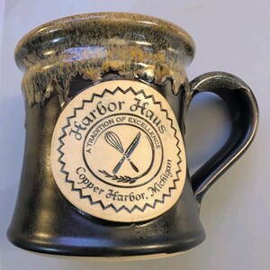 May include: A ceramic mug with a black base and a tan rim featuring a drip glaze effect. A circular tan medallion on the front displays the text Harbor Haus, A Tradition of Excellence, Copper Harbor, Michigan, with a whisk and knife graphic.