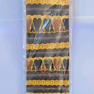 May include: A black necktie featuring a repeating pattern of gold and brown heart-shaped motifs and intricate gold filigree bands. The tie is encased in a clear plastic sleeve and displays the text Claudio Riva and MADE IN ITALY on the fabric.