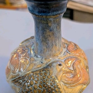 May include: A small ceramic vase with a narrow neck and a bulbous body. The neck is glazed in a deep blue, while the body features a relief design of a bird with textured wings, finished in shades of tan, peach, and blue with a glossy glaze.