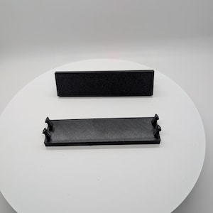 May include: Two black rectangular objects are displayed on a white surface. One is a solid block, and the other is a tray-like shape with small protrusions at each end. The objects appear to be 3D-printed.