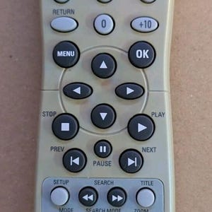 May include: A light gray Magnavox remote control featuring a red power button at the top, a numeric keypad, directional navigation arrows, and playback control buttons including play, pause, stop, skip, and search functions labeled with text.