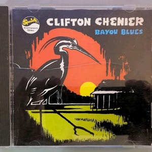 May include: A music CD case featuring the album Bayou Blues by Clifton Chenier. The cover art displays a black silhouette of a heron standing in a green marsh against a bright orange sunset and a yellow sun, with a small wooden shack on the right.