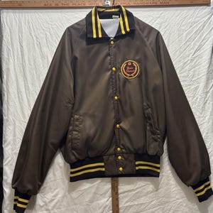May include: A brown satin-finish jacket with yellow striped ribbed cuffs, collar, and hem. It features yellow snap buttons and a circular patch on the left chest with the text "Master Mechanic" inside a laurel wreath design.