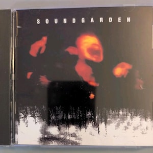 May include: A Soundgarden music CD in a clear plastic jewel case. The cover art features a dark background with glowing orange, abstract shapes above a black and white silhouette of a forest reflected in a snowy or icy landscape at the bottom.