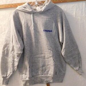 May include: A heather gray hooded sweatshirt featuring long sleeves, a front kangaroo pocket, and white drawstring cords. The word Graybar is printed in blue text on the upper left chest area. The garment is displayed against a plain background.