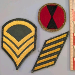 May include: Three embroidered military patches next to a ruler showing inches. One is a shield-shaped dark green patch with yellow chevrons, one is a circular red patch with a black hourglass, and one is a diagonal dark green patch with yellow stripes.