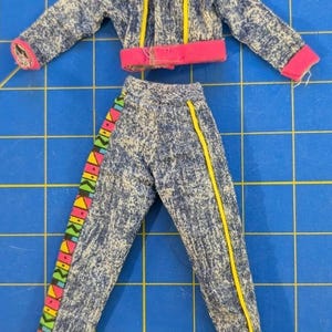May include: A doll-sized jacket and pants set made of blue acid-wash fabric. The jacket features a pink collar and hem, with a geometric pattern in pink, yellow, and green across the shoulders. The pants have a matching geometric side stripe.