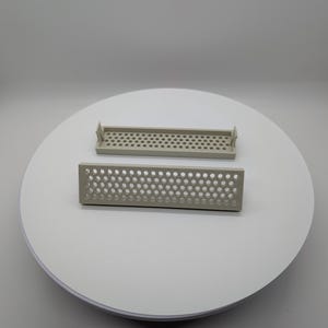 May include: Two rectangular, light gray plastic ventilation grilles with a grid of small, evenly spaced holes. One grille is positioned above the other, both on a white circular surface.