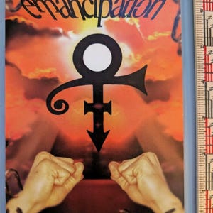 May include: A rectangular print featuring the word Emancipation in black script at the top, a large black symbol centered over a sunset background, and two clenched fists at the bottom with broken chains. A ruler shows a height of 6.5 inches.
