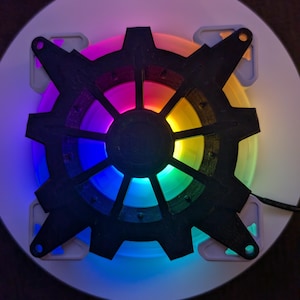 May include: A black, gear-shaped computer fan with vibrant, multicolored LED lights. The fan is mounted on a white circular base with mounting tabs. The lights cycle through a rainbow of colors, creating a visually striking effect.