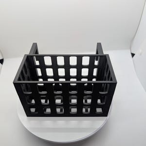 May include: A black, rectangular basket with a grid-like pattern of square cutouts. The basket has two vertical handles and sits on a white, circular base. The design is modern and minimalist, suitable for storage or display.