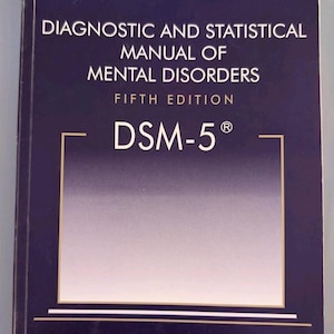 May include: A dark blue hardcover book titled Diagnostic and Statistical Manual of Mental Disorders Fifth Edition DSM-5 with white text and a gold-outlined rectangular frame on the front cover, published by the American Psychiatric Association.