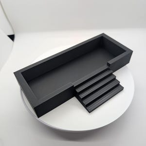 May include: A black plastic rectangular tray with a raised platform on one side. The platform has four steps leading up to the tray.