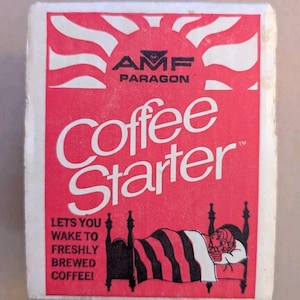 May include: A rectangular white and red box labeled AMF PARAGON Coffee Starter. The design features a stylized sun, a graphic of a person sleeping in a bed under a striped blanket, and the text LETS YOU WAKE TO FRESHLY BREWED COFFEE!
