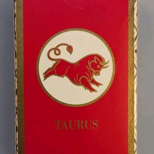 May include: A rectangular deck of playing cards box featuring a vibrant red background with a gold border. A central white circle contains a red bull illustration. The word TAURUS is printed in gold below the circle. The top edge shows U.S. PLAYING.