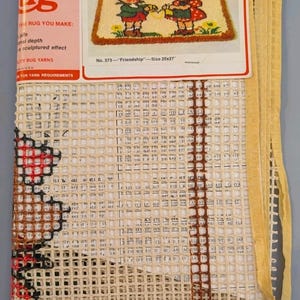 May include: A latch hook rug kit featuring a printed label with the text No. 373 Friendship Size 20x27. The kit includes a grid canvas with partial red and brown yarn stitching, a pink header, and an illustration of two children holding hands.