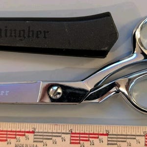 May include: Silver-colored metal Gingher scissors with a black protective sheath. The blade is engraved with the word Gingher. A ruler is positioned below the scissors, showing the tool measures approximately 8 inches in total length.