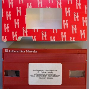 May include: A red VHS tape labeled Lutheran Hour Ministries with a white sticker reading An important message from Dr. Dale A. Meyer with preview scenes from RED BOOTS FOR CHRISTMAS Television Special, paired with a matching red cardboard sleeve.