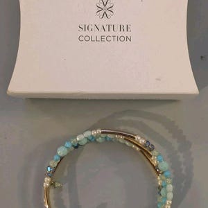 May include: A multi-strand wrap bracelet featuring gold-tone metal tubes interspersed with small beads in shades of light blue, turquoise, and white, positioned below a white gift box labeled Signature Collection.