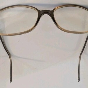 May include: A pair of eyeglasses with a translucent brown, oval-shaped plastic frame and clear lenses. The frame features small silver-toned metal rivets at the outer corners where the temples attach to the front, with thin, curved temple arms.