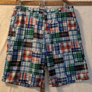 May include: Multi-colored plaid shorts featuring a patchwork design of blue, green, red, and white squares. The shorts have a waistband with belt loops and a button closure. A wooden ruler is visible at the top, showing measurements in inches.