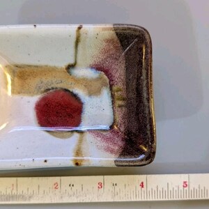 May include: A rectangular ceramic dish with a white base, featuring a prominent red circle, brown accents, and dark edges. A white ruler is positioned along the bottom and right side, showing the dish is approximately 4.5 inches wide and 3 inches tall.