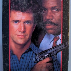 May include: A VHS tape cover for the film Lethal Weapon 2 featuring Mel Gibson and Danny Glover. The cover displays the movie title in white and red text, a black handgun, and a blue Warner Bros 75th anniversary promotional sticker at the bottom.