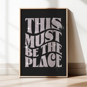 Puede incluir: Un póster enmarcado con la frase "THIS MUST BE THE PLACE" en letras plateadas brillantes sobre un fondo negro. El póster tiene un marco de madera clara y detalles de estrellas.