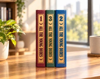 May include: Three hardcover books titled All The Young Dudes, numbered 1, 3, and 2 on their spines. The books are bound in red, green, and blue covers with gold lettering and crescent moon symbols, standing upright in a row on a wooden surface.