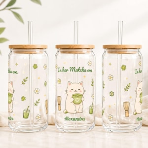Op de afbeelding: Drie doorzichtige glazen bekers met bamboe deksels en glazen rietjes. Elke beker is voorzien van een cartoonkat met een matchakom, groene bloemmotieven, kleine sterretjes en de tekst In her Matcha era met de naam Alexandra in het groen.