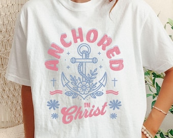 May include: White t-shirt with the text "ANCHORED IN CHRIST" in pink and blue. The design features an anchor, flowers, and crosses. The shirt is a casual, oversized fit.