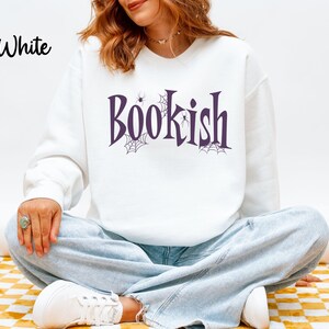 May include: A white long-sleeve crewneck sweatshirt featuring the word Bookish printed in a dark purple serif font. The design includes small black spider web illustrations and tiny spiders integrated into the lettering on the front chest.